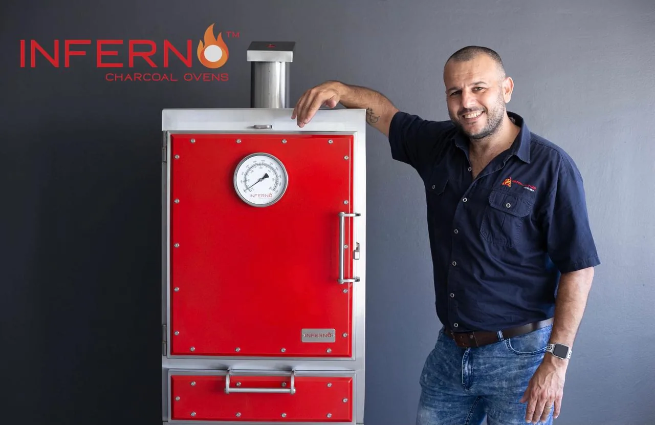 José Luis Gomez standing beside a red Inferno charcoal oven, one arm resting on its top, against a dark grey wall.