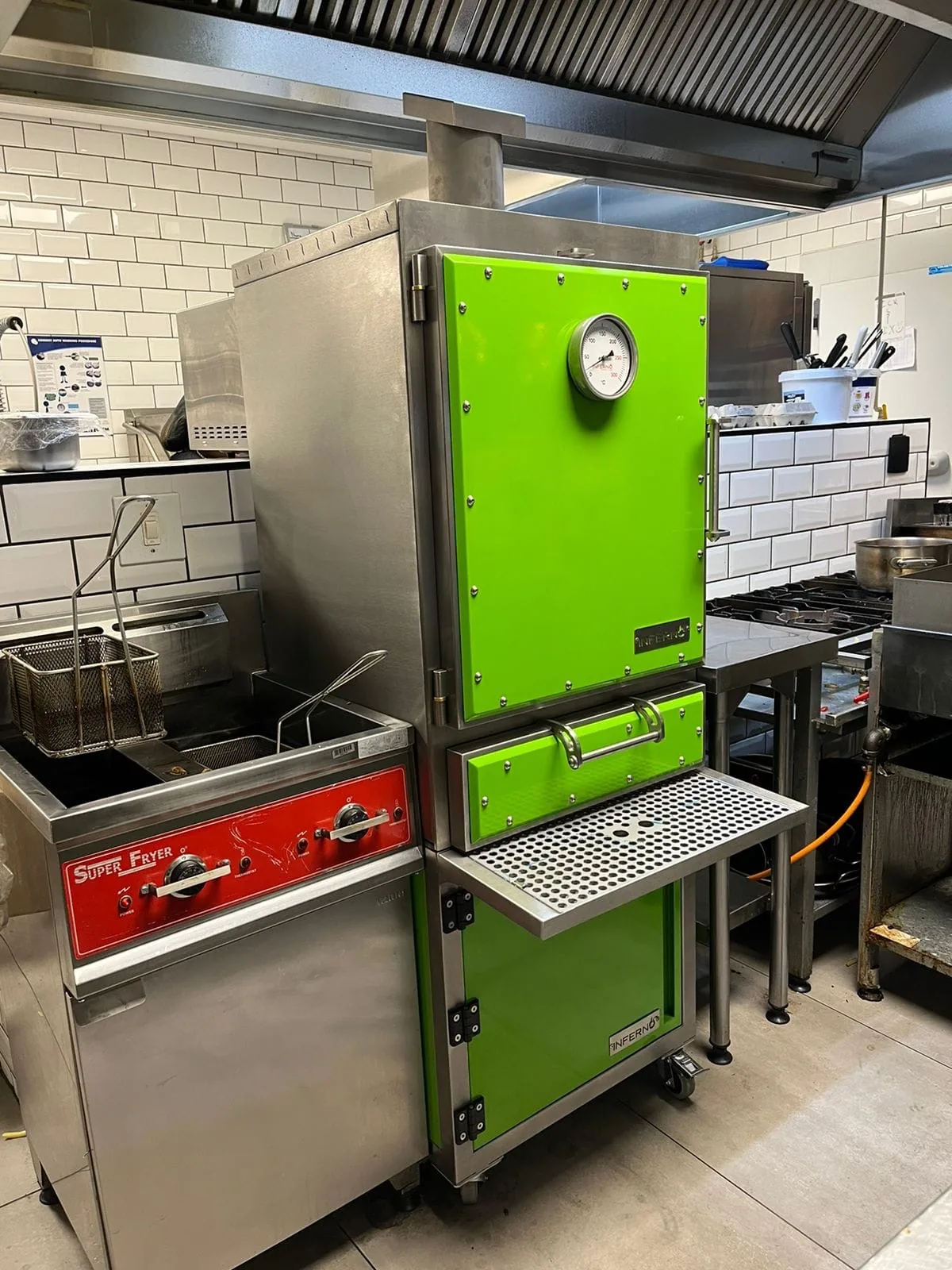 A green Inferno charcoal oven on a commercial cooking line, beside a fryer.