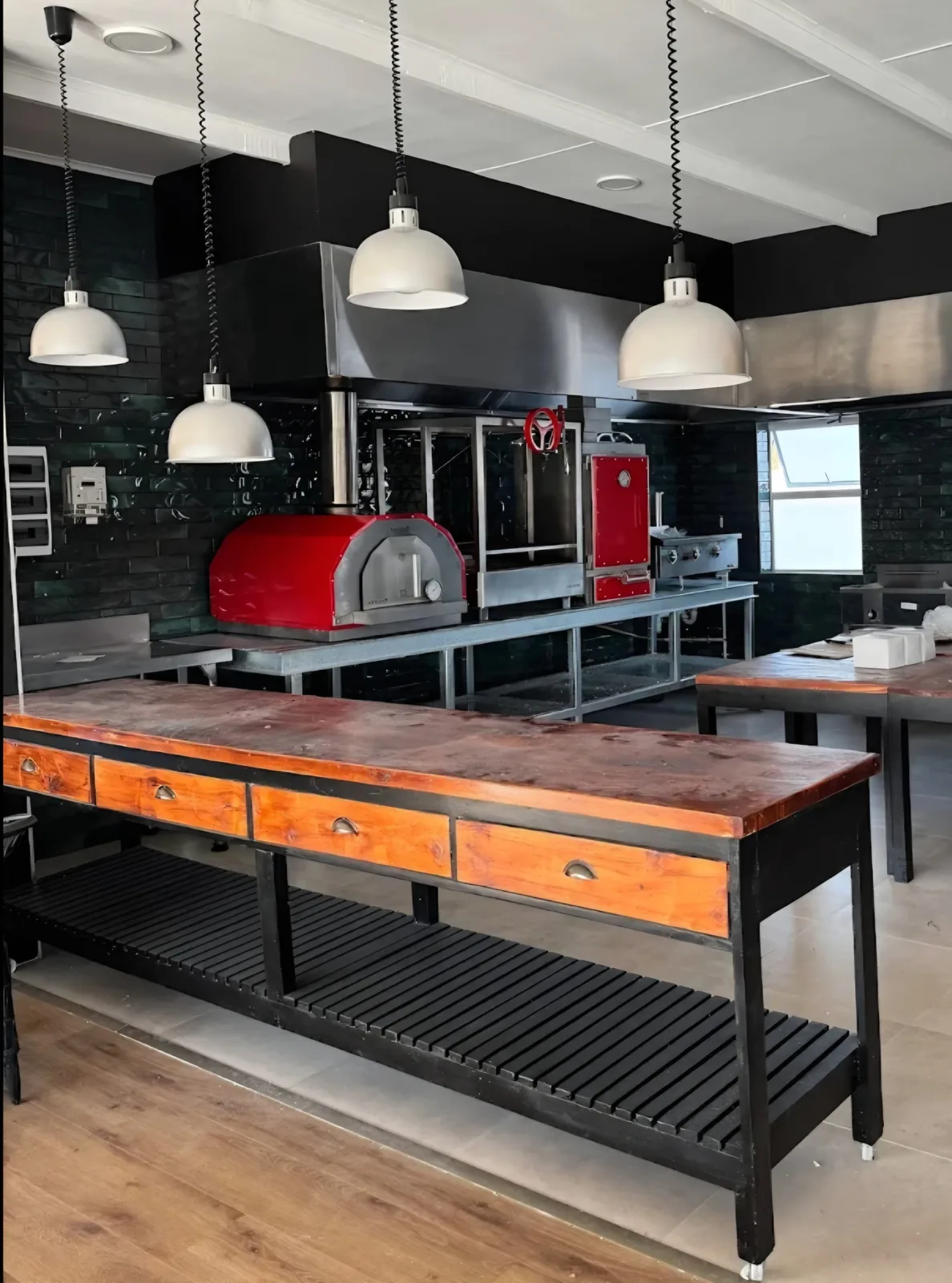 A red Inferno charcoal oven and a red pizza oven in an open kitchen with pendant lights and a long timber table.