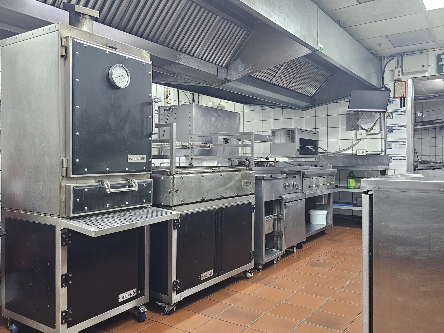 A black Inferno charcoal oven at the head of a long commercial cooking line, beside a grill.