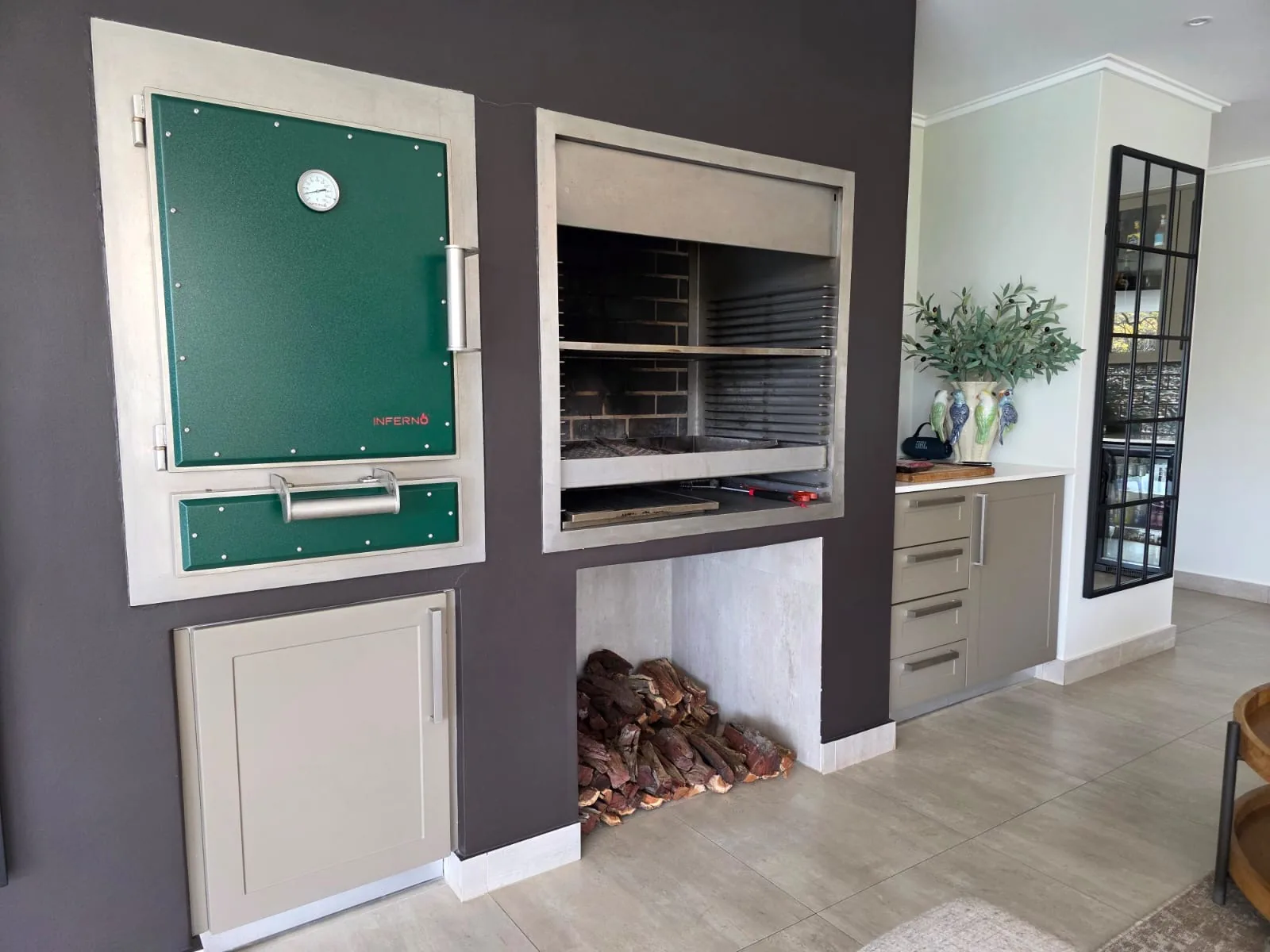 A green Inferno charcoal oven built into a feature wall beside a built-in braai, with firewood stacked below.