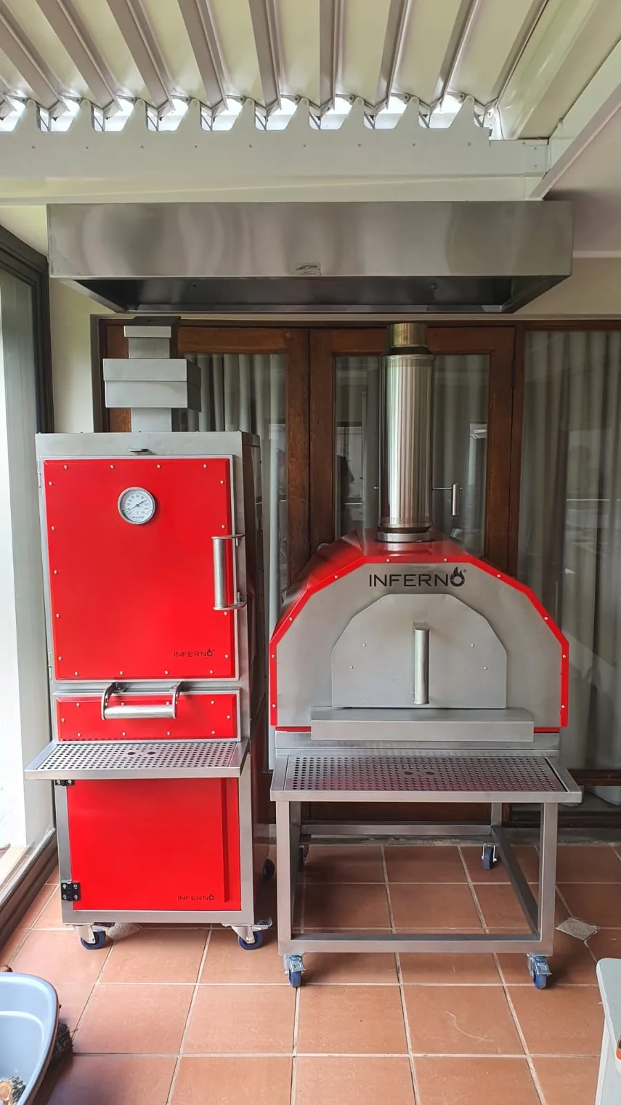 A red Inferno charcoal oven beside an Inferno pizza oven on a covered terrace with a terracotta floor.