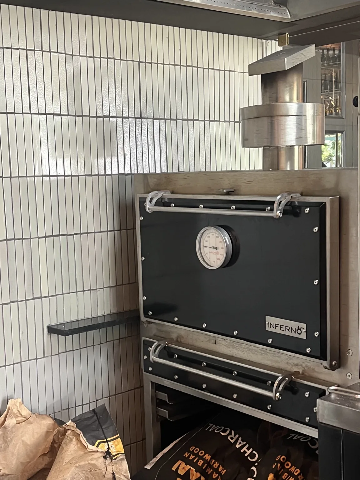 A black Inferno charcoal oven built into a tiled wall, its chimney rising above it.