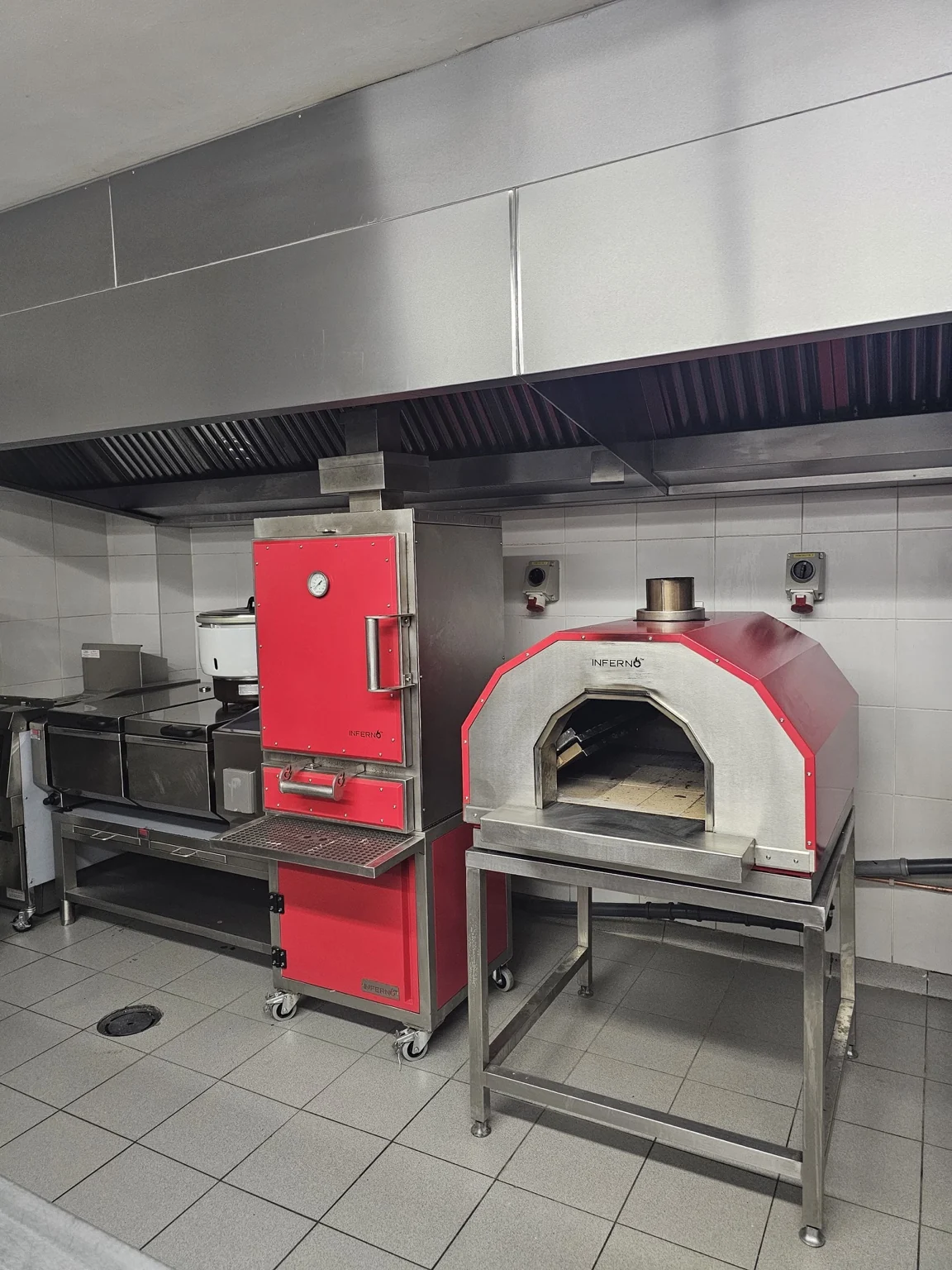 A red Inferno Alto installed in a tiled commercial kitchen under an extraction canopy, beside an Inferno X Pizza Oven on its stand.